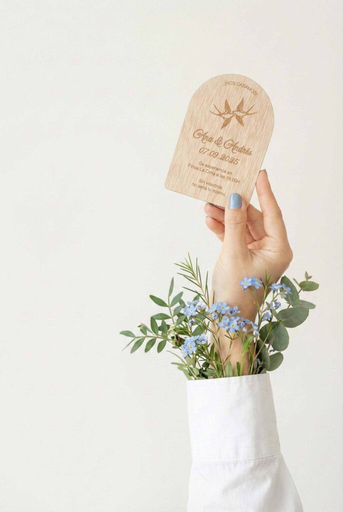 Invitación de boda de madera en forma de arco sujeta con la mano entre flores no me olvides