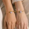 Dos pulseras No me olvides unidas por imu00e1n en las muu00f1ecas de dos personas, con flor azul y cadena dorada.