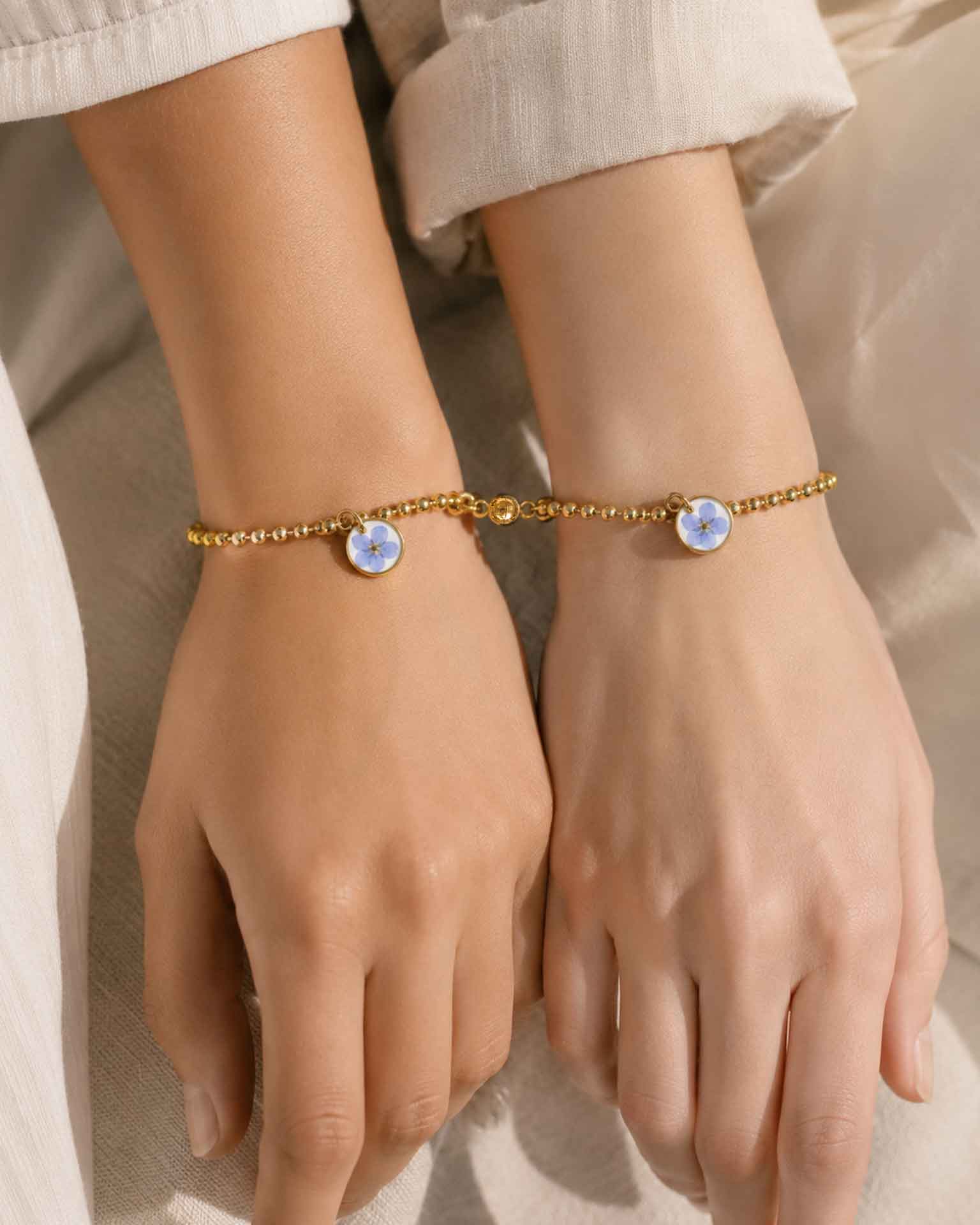 Dos pulseras No me olvides unidas por imán en las muñecas de dos personas, con flor azul y cadena dorada.
