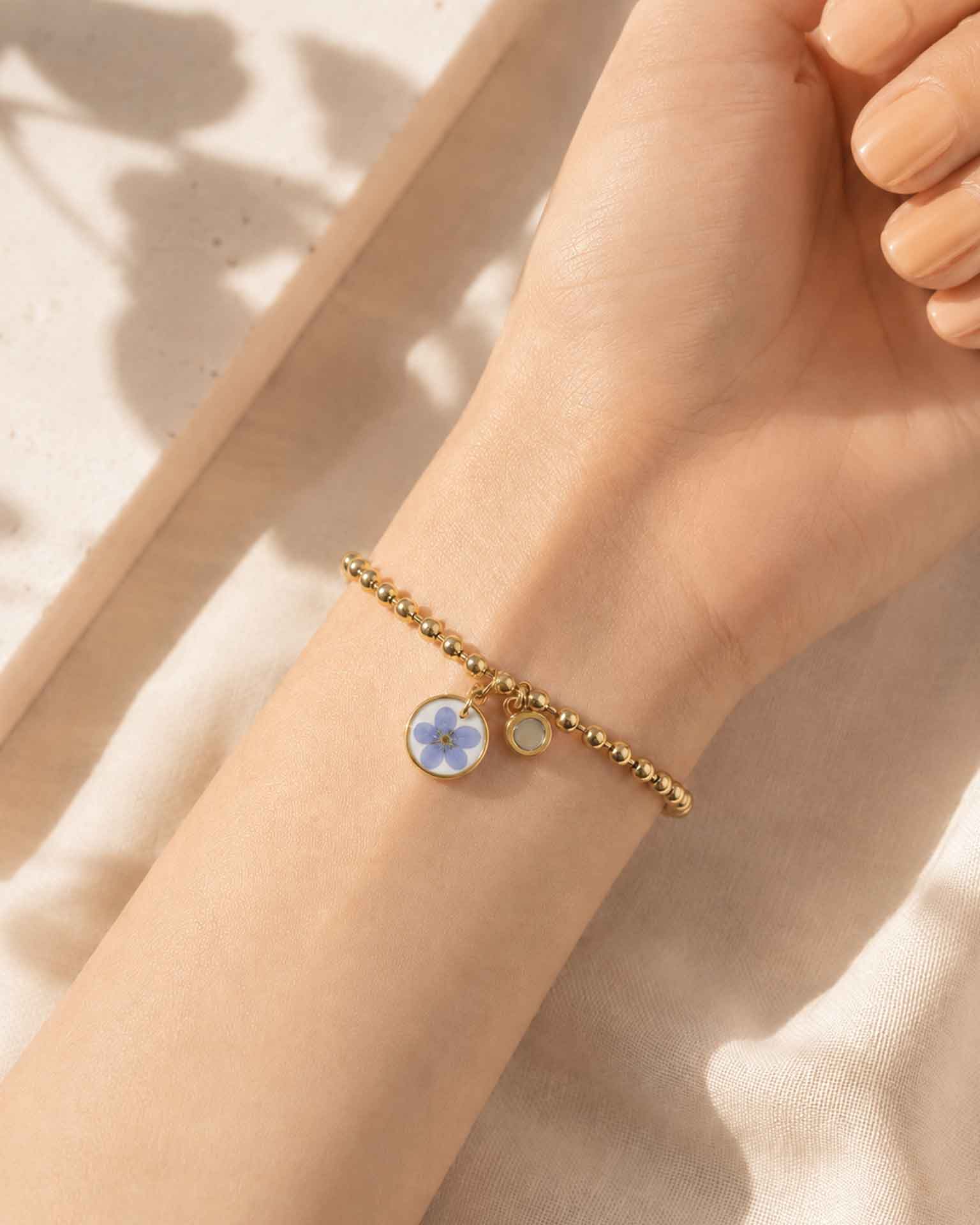 Pulsera No me olvides dorada en la muu00f1eca con charm de flor azul y detalle imantado.