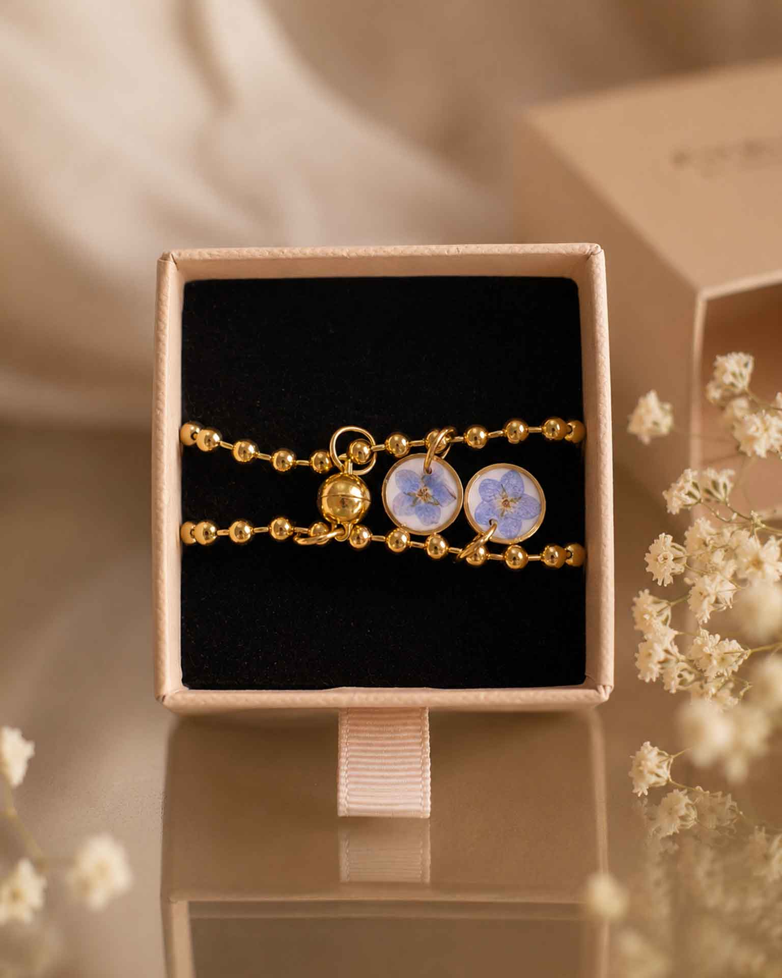 Pulseras No me olvides doradas presentadas en caja de regalo con flores azules y pieza imantada central.