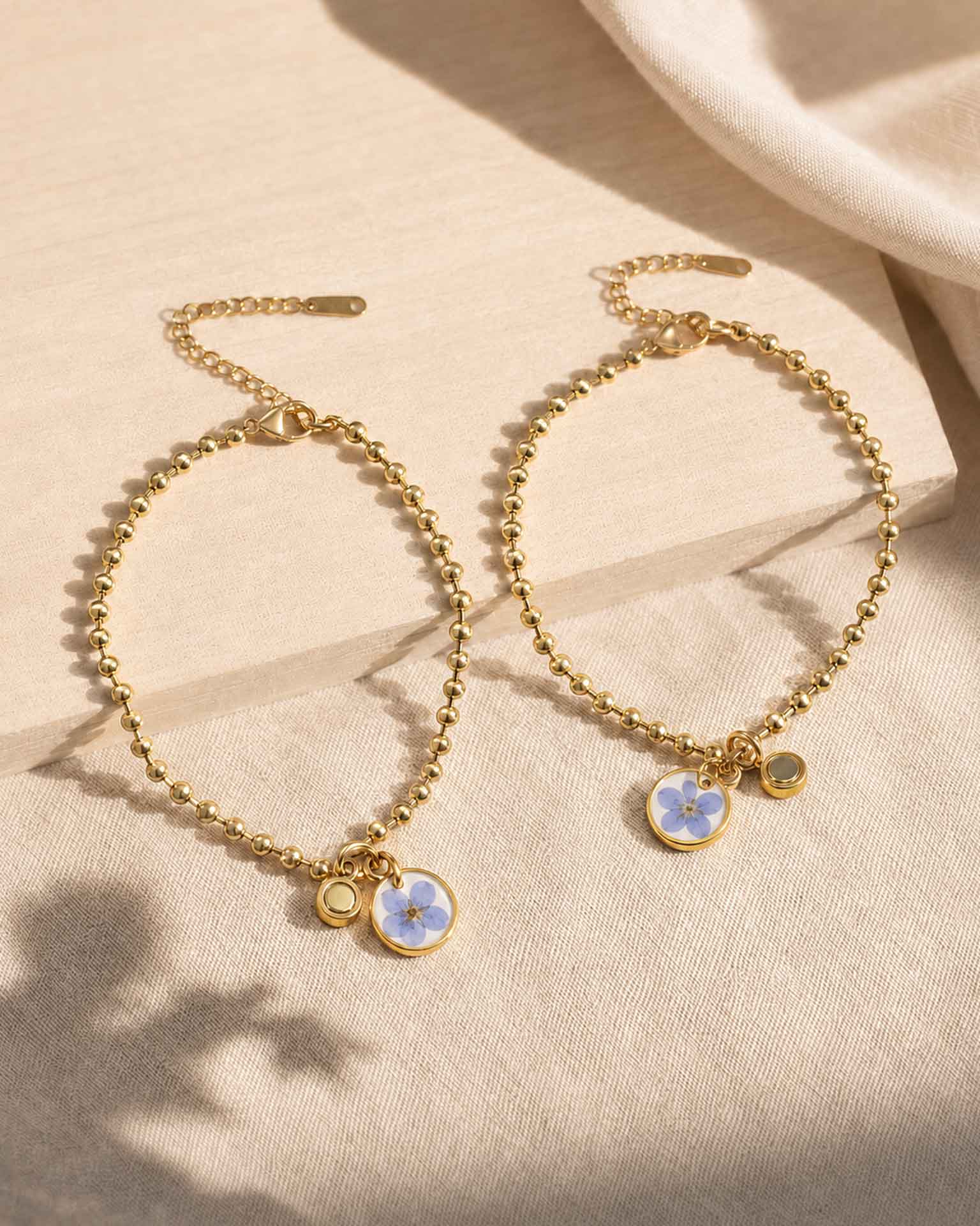 Dos pulseras No me olvides doradas con charms de flor azul y piezas imantadas para compartir.
